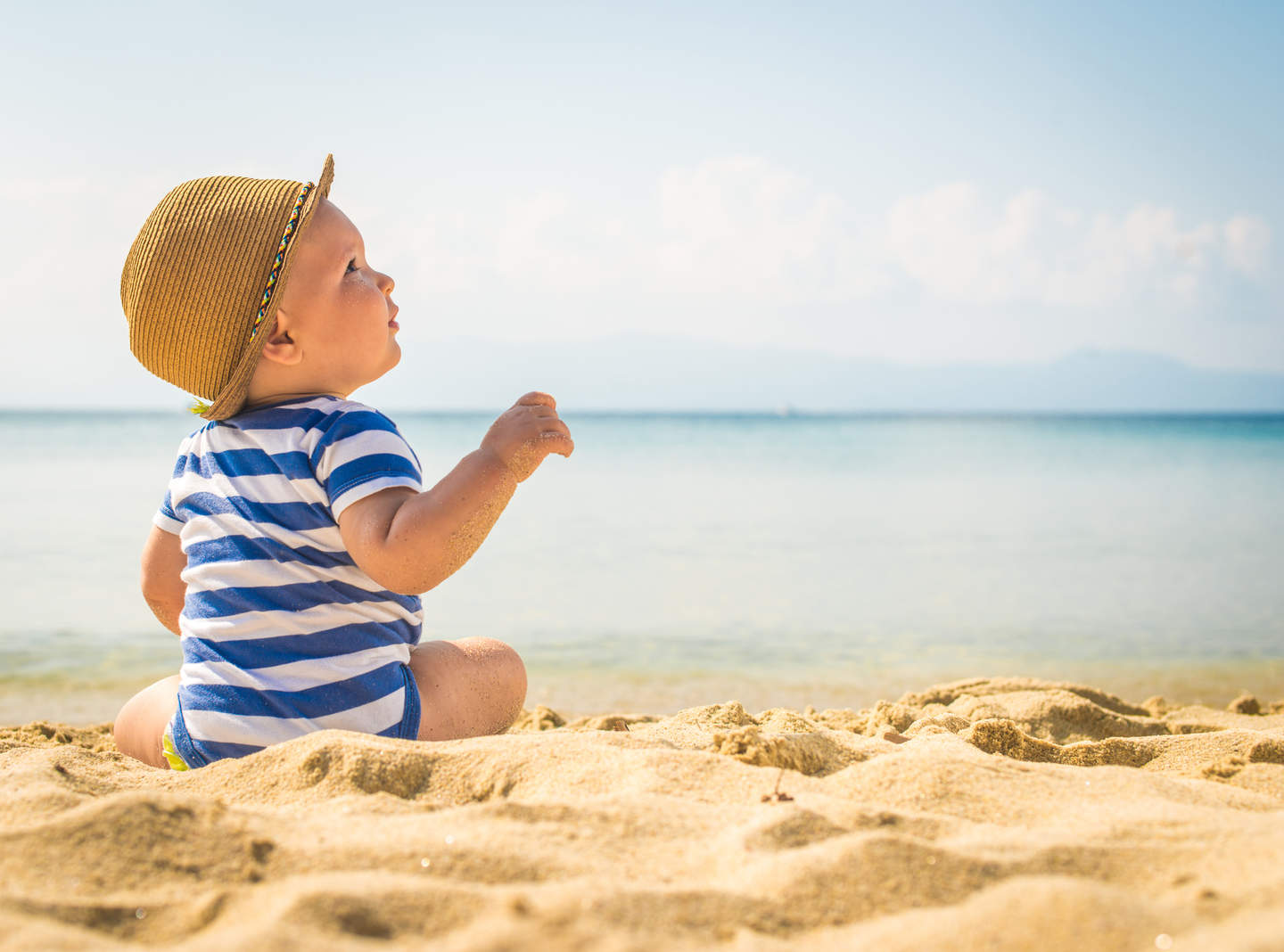 Baby in hat and blue striped romper sitting on beach looking up