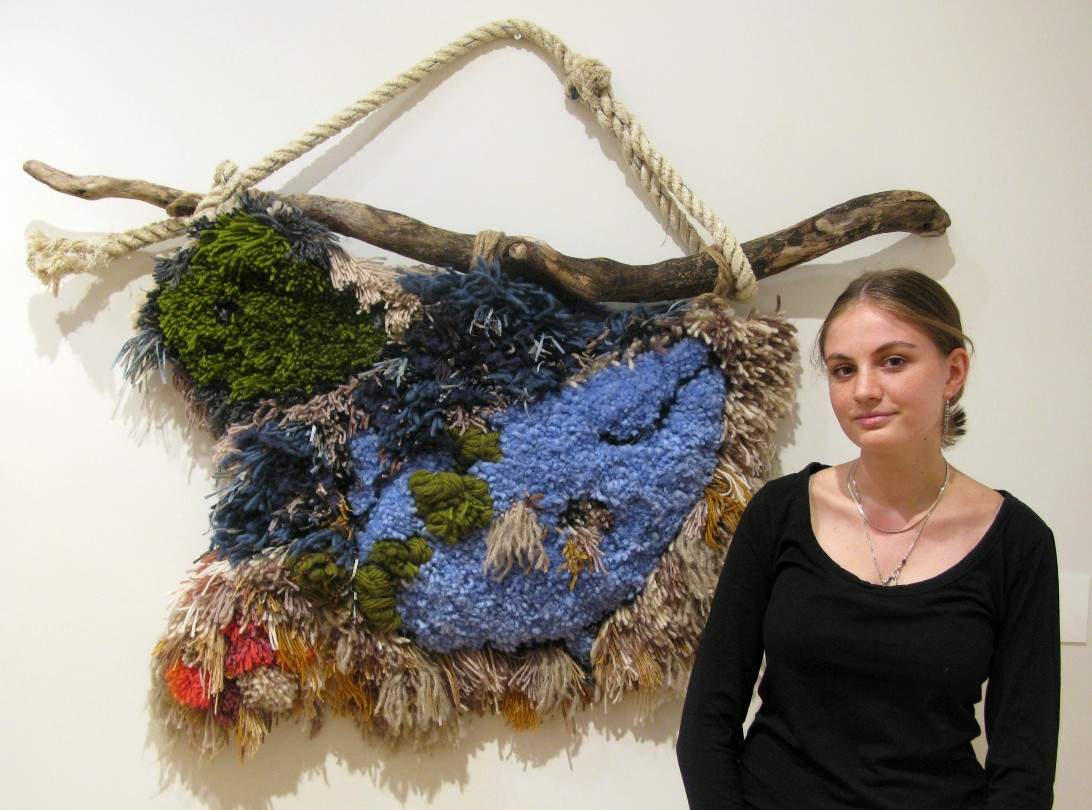 A young girl stands in front of an artwork made of wool suspended by a long branch and rope.