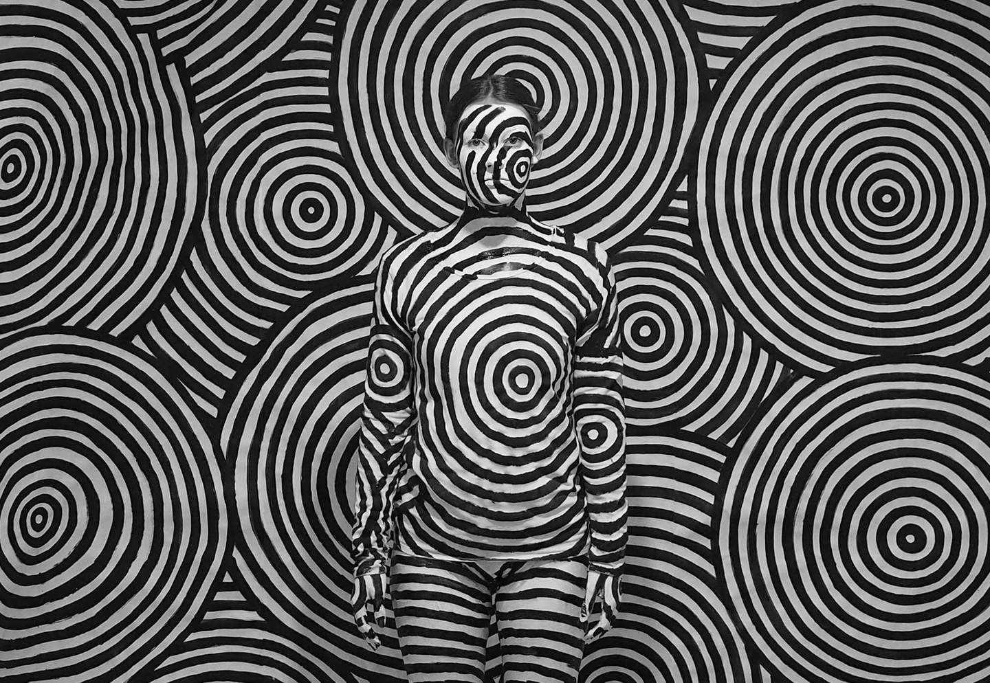 A young person is painted in black and white swirls camouflaged against the same patterned backdrop. 