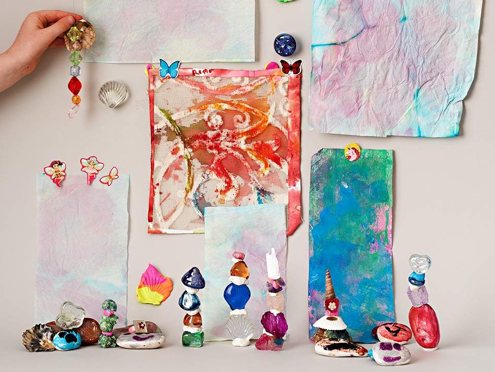A display of stacked colorful stones and small paintings on paper on wall, a person's hand holds up a trinket.