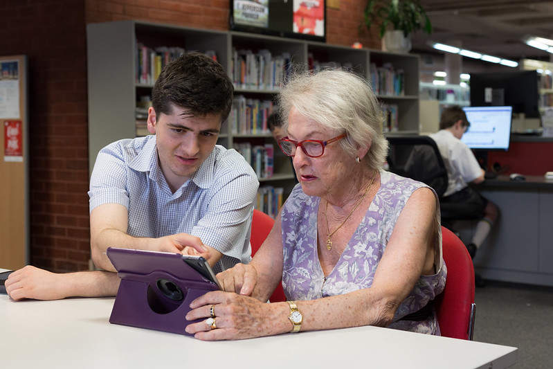 Young man showing an older woman how to use her iPad device at the library as part of a volunteer program