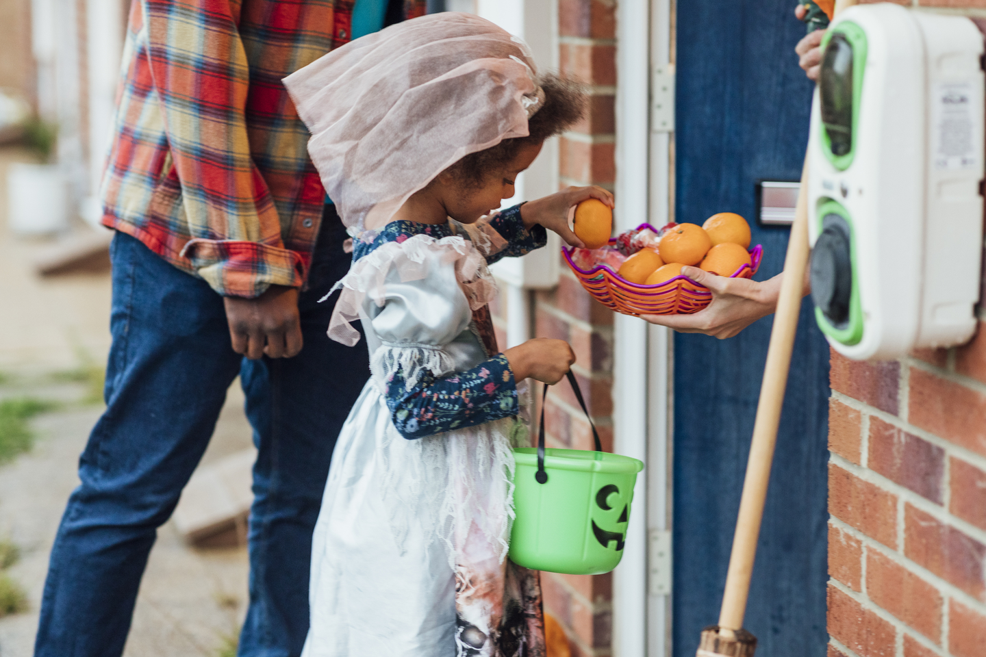 A low angle close up side view of a little girl choosing a healthy treat to put in her green trick or treat bucket. She is being accompanied by her father as they knock on the neighbours doors on Halloween.