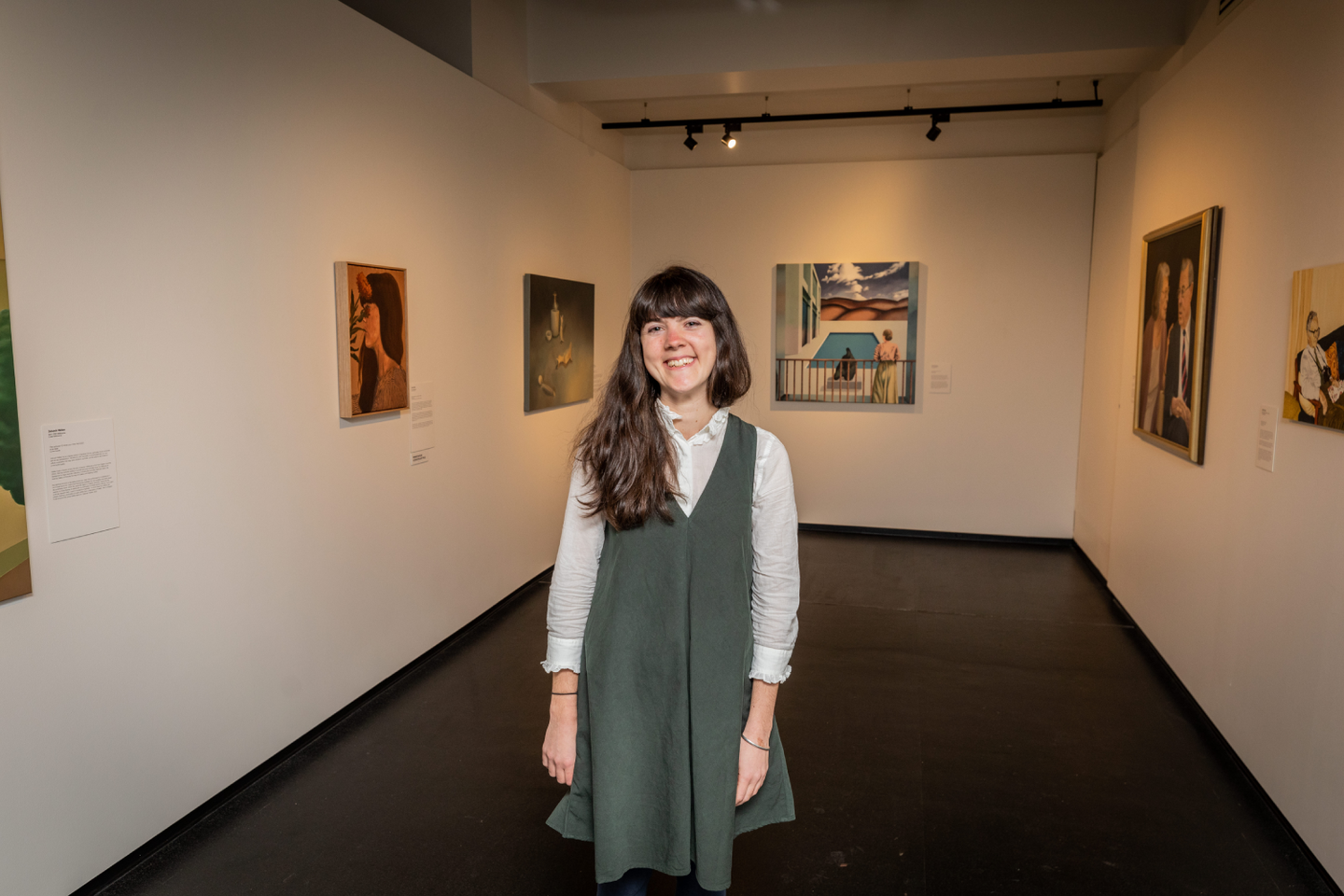 Woman with brown hair smiling in front of gallery filled with paintings