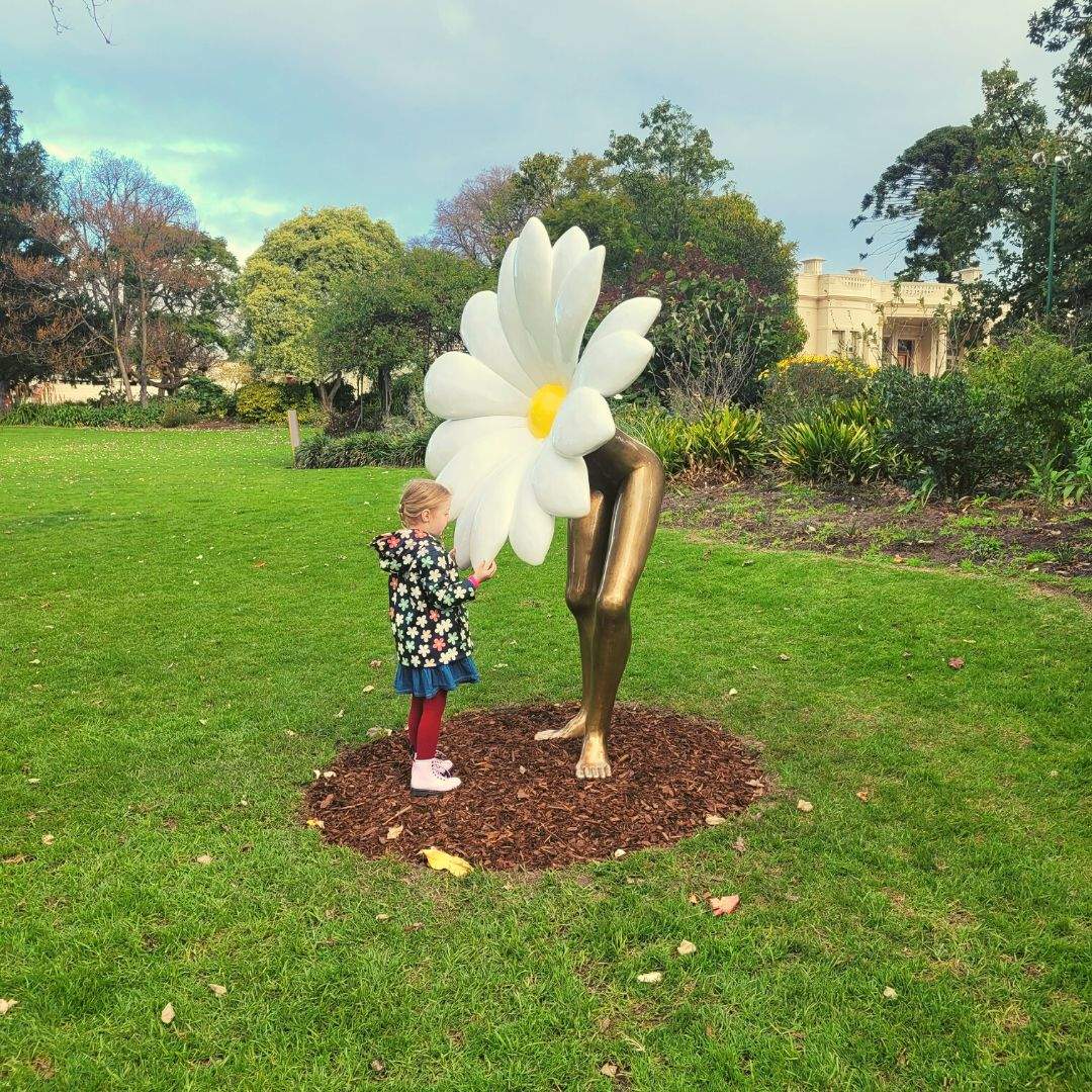 Littel girl in a raincoat touching a daisy sculpture with bronze legs.