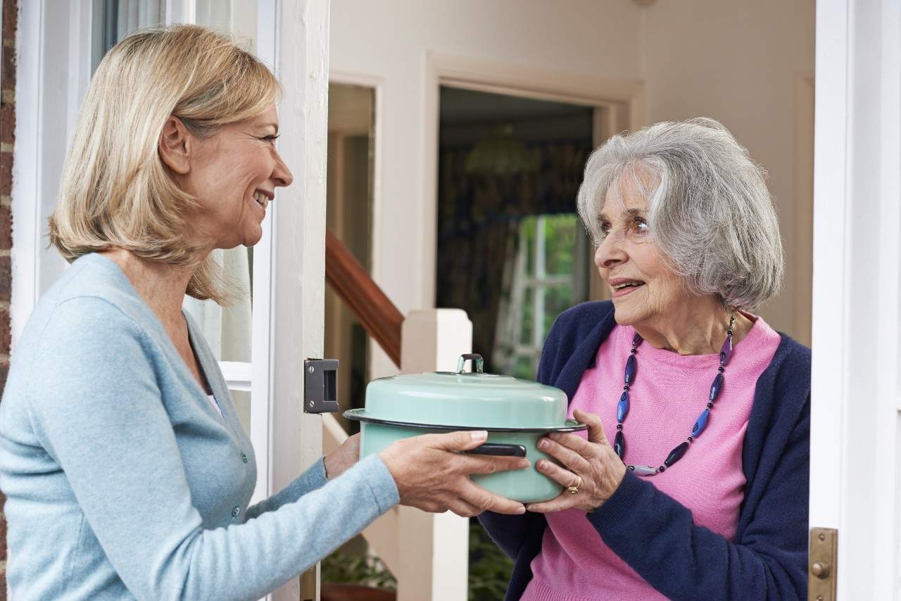 A woman smiling and holding a casserole dish passing it to an older woman in a doorway. 