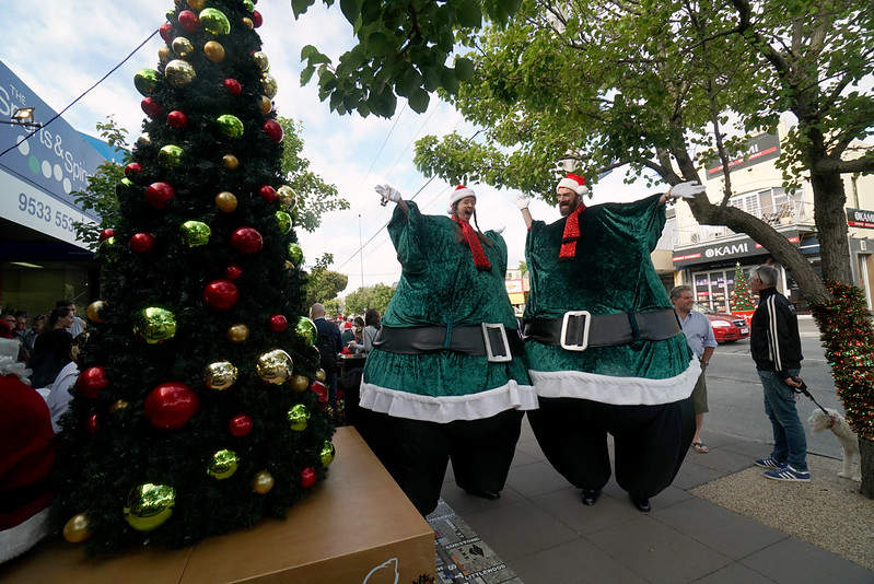 entertainers on stitlts dressed in green Santa outfits beside a Christmas tree on a Bayside shopping strip