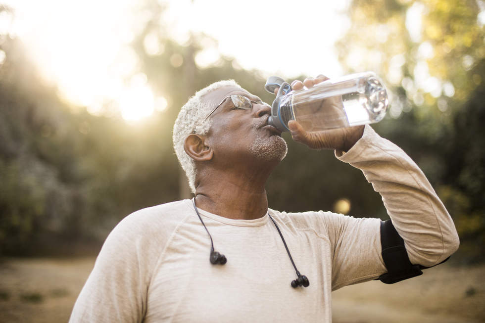 Older man, with earbuds and exercise mobile phone arm strap,, standing outside drinking water from a bottle