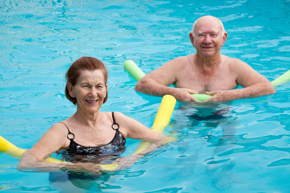 Older woman and man swimming with pool noodles in warm water pool