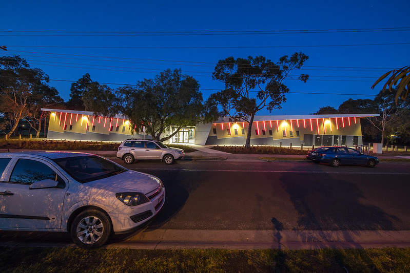 Sports pavilion building, lit up at night with trees, cars and footpath