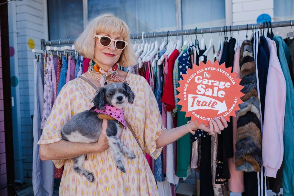 Woman holding a small dog and a Garage Sale Trail sign in front of a rack of clothes