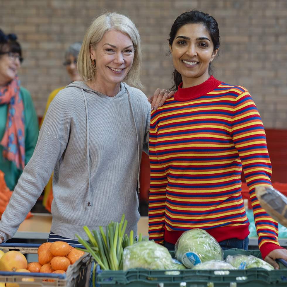 Two smiling women standing behind a table with food in trays sitting on top of it in a food relief centre