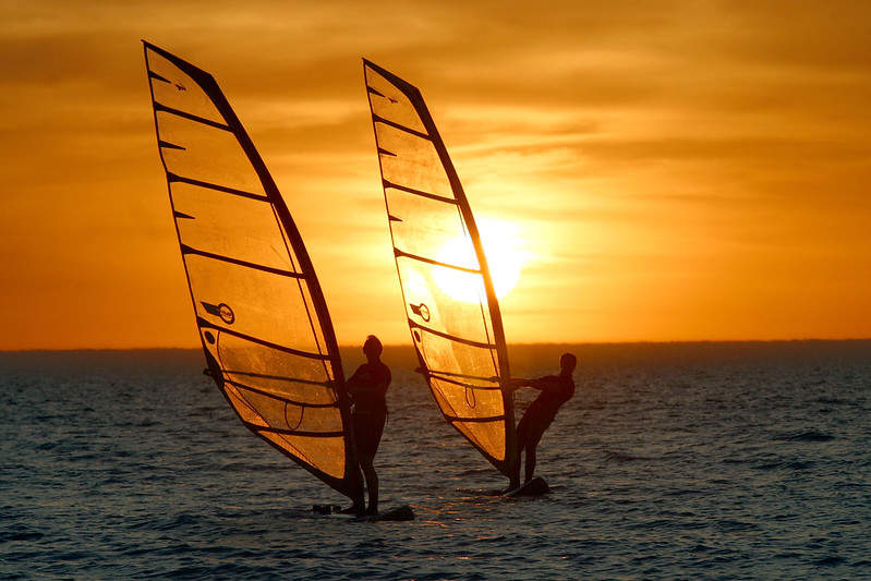 two people windsurfing out on the open water with a yellow and orange sunset in the background