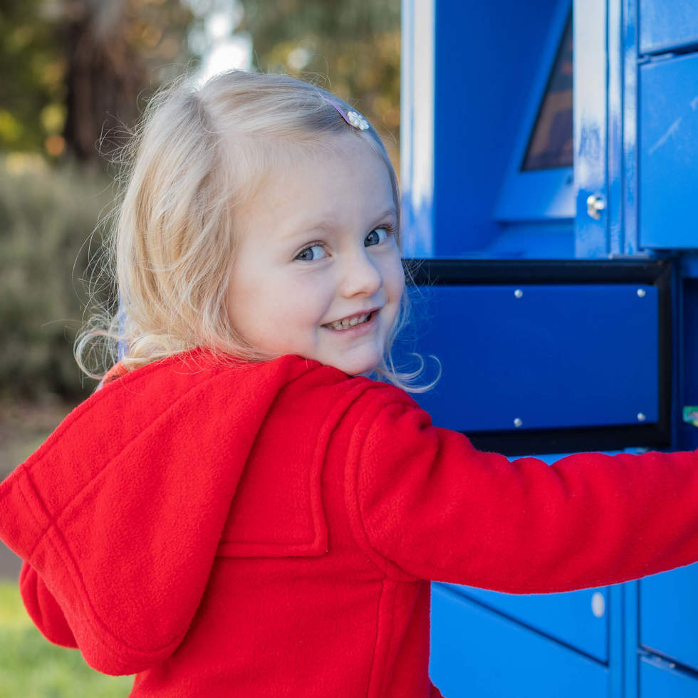 Little girl with blonde hair in a bright red coat collecting books from the Hurlingham Park Library Locker