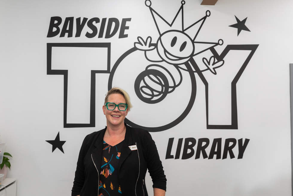 The Manager of the Toy Library stands smiling proudly in front of a sign that reads Bayside Toy Library