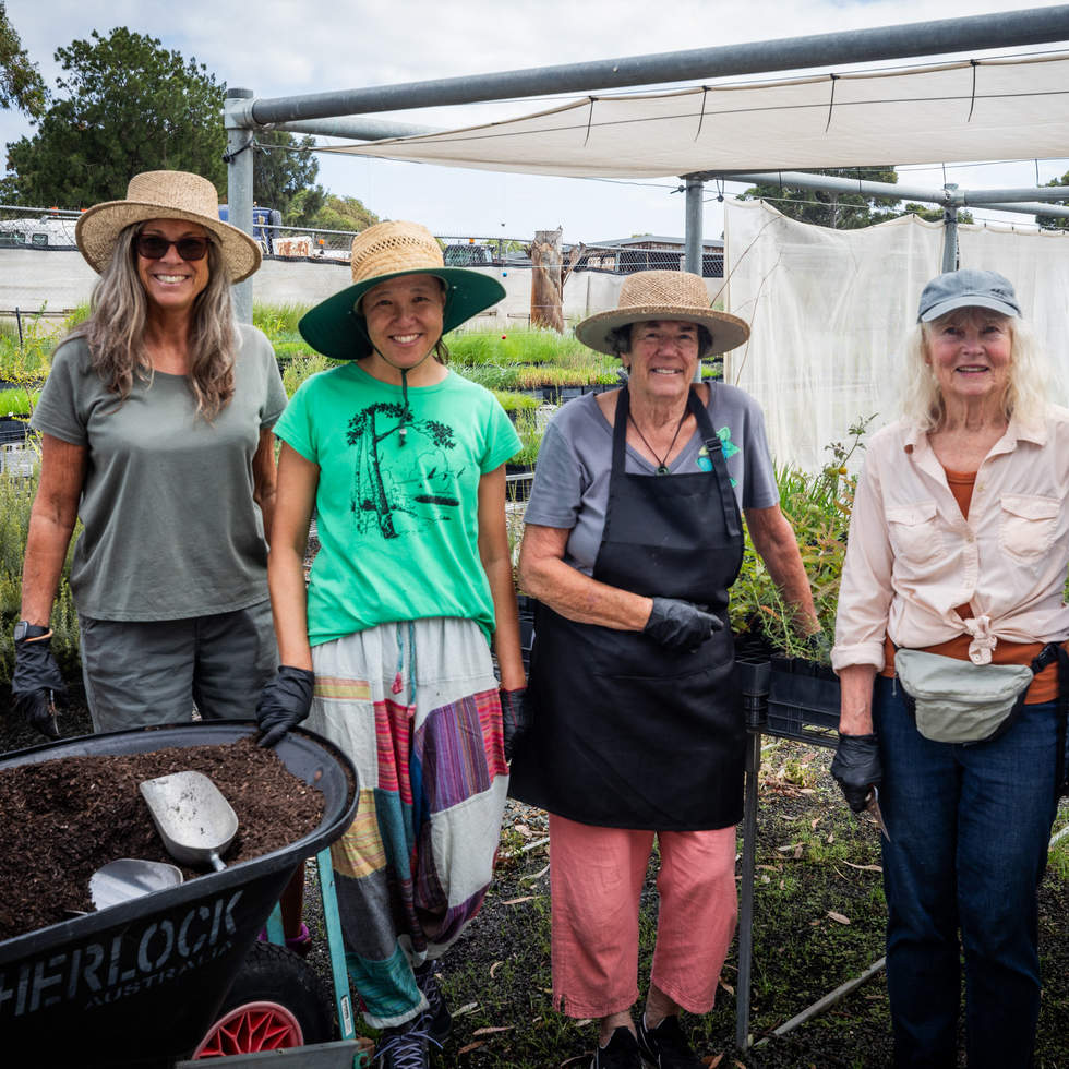 Five people smiling at the camera in front of plants and wheelbarrow