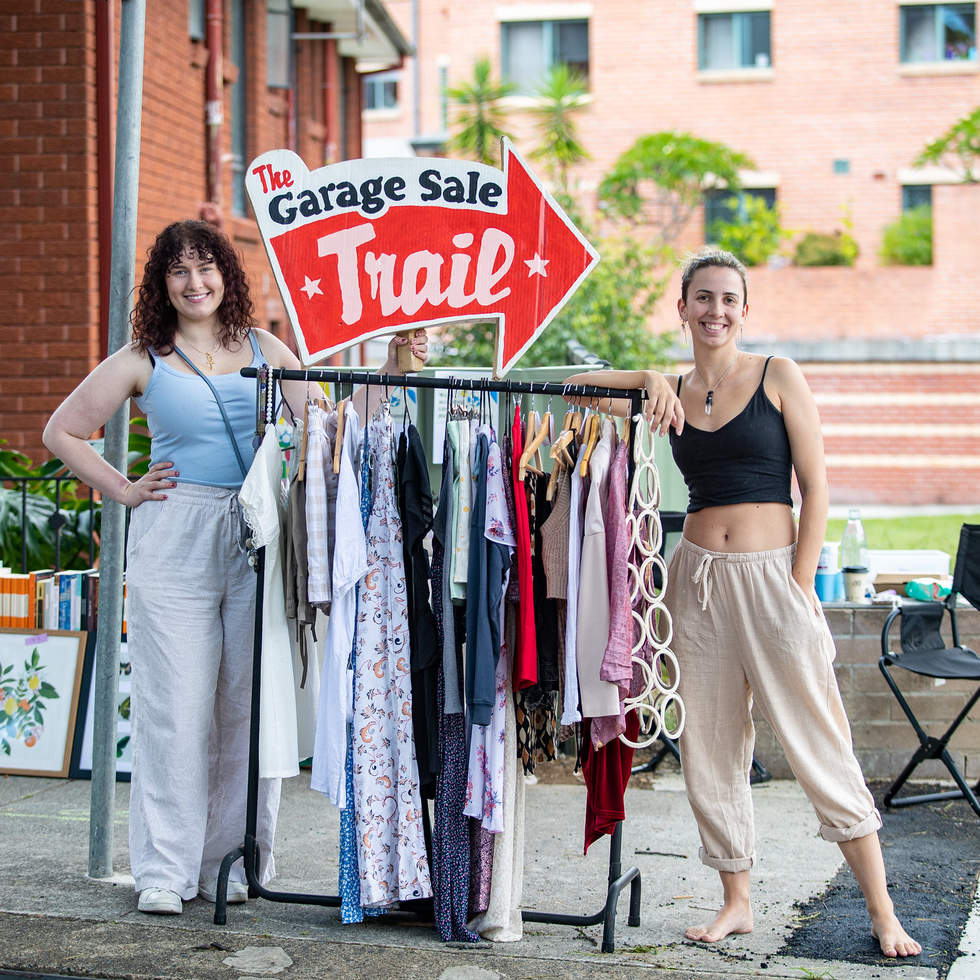 Two women stand either side of a clothing rack in a parking space, with a red arrow sign above the clothing rack saying 'The Garage Sale Trail'